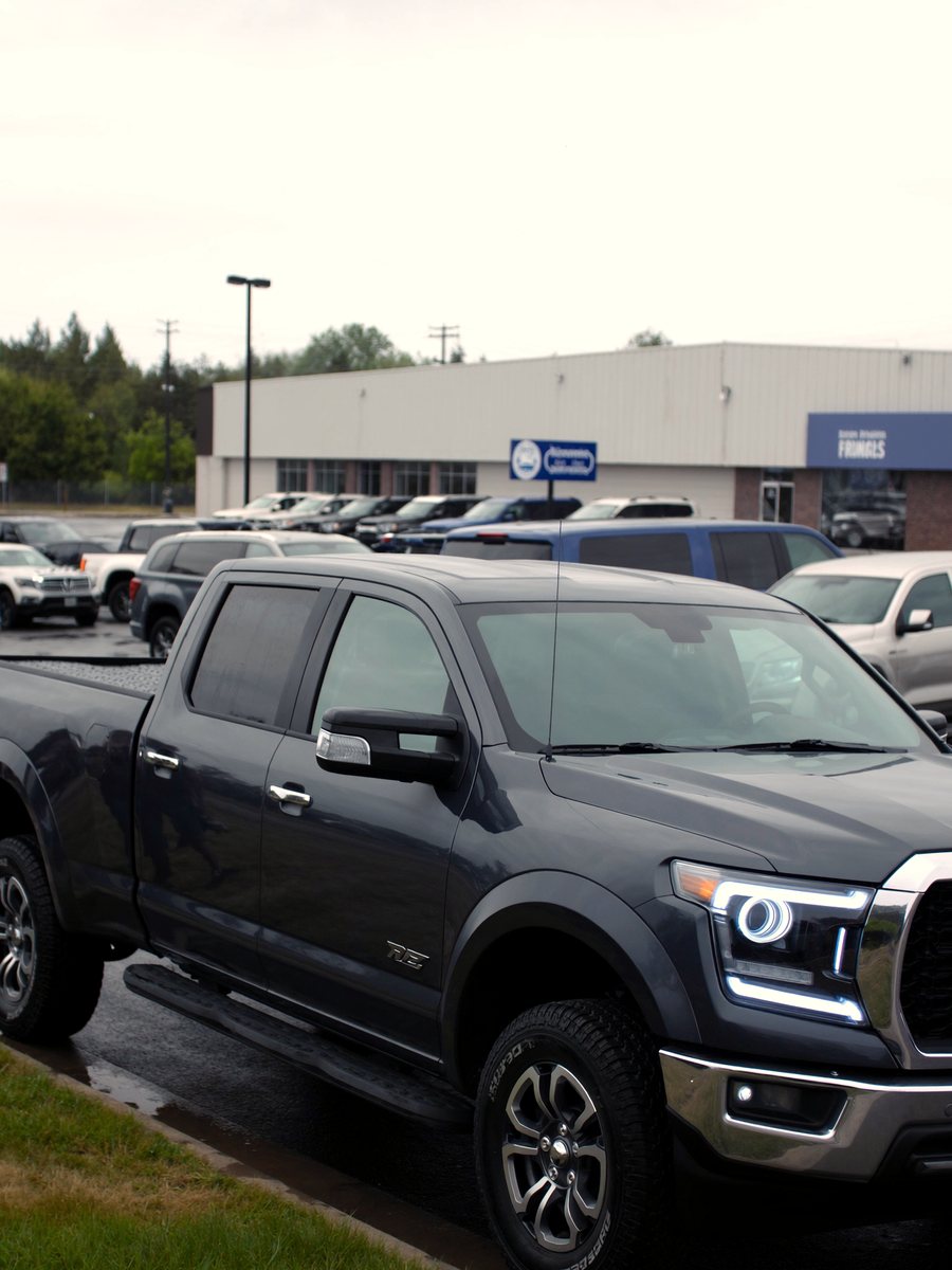 A dark grey crew cab pickup on wet asphalt after rain, rows of new trucks behind it under overcast sky.