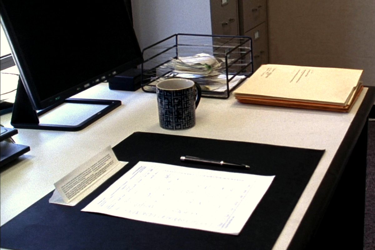 A printed order form and pen on a desk blotter, with a coffee mug, a wire in-tray and a stack of manila folders behind.