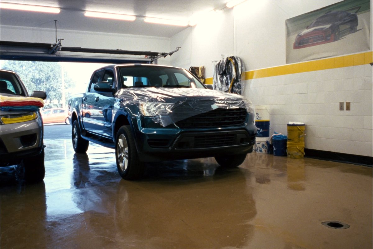 A new crew cab pickup on the wet floor of a dealership prep bay, shipping film still across its hood, buckets along the wall.